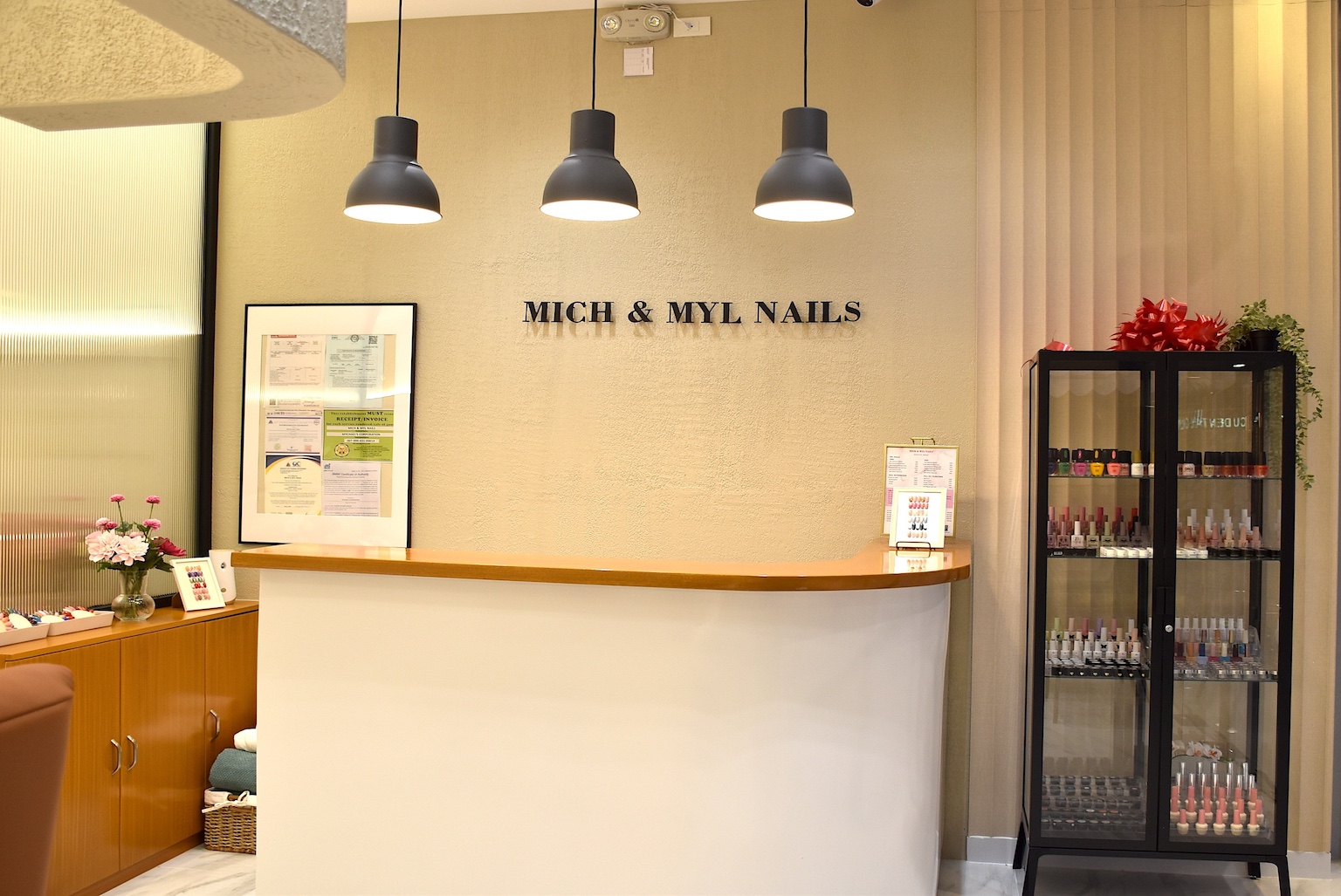 Reception desk at a nail salon with 'MICH & MYL NAILS' on the wall, glass display cabinet of nail polishes to the right, and a small flower arrangement on the left.
