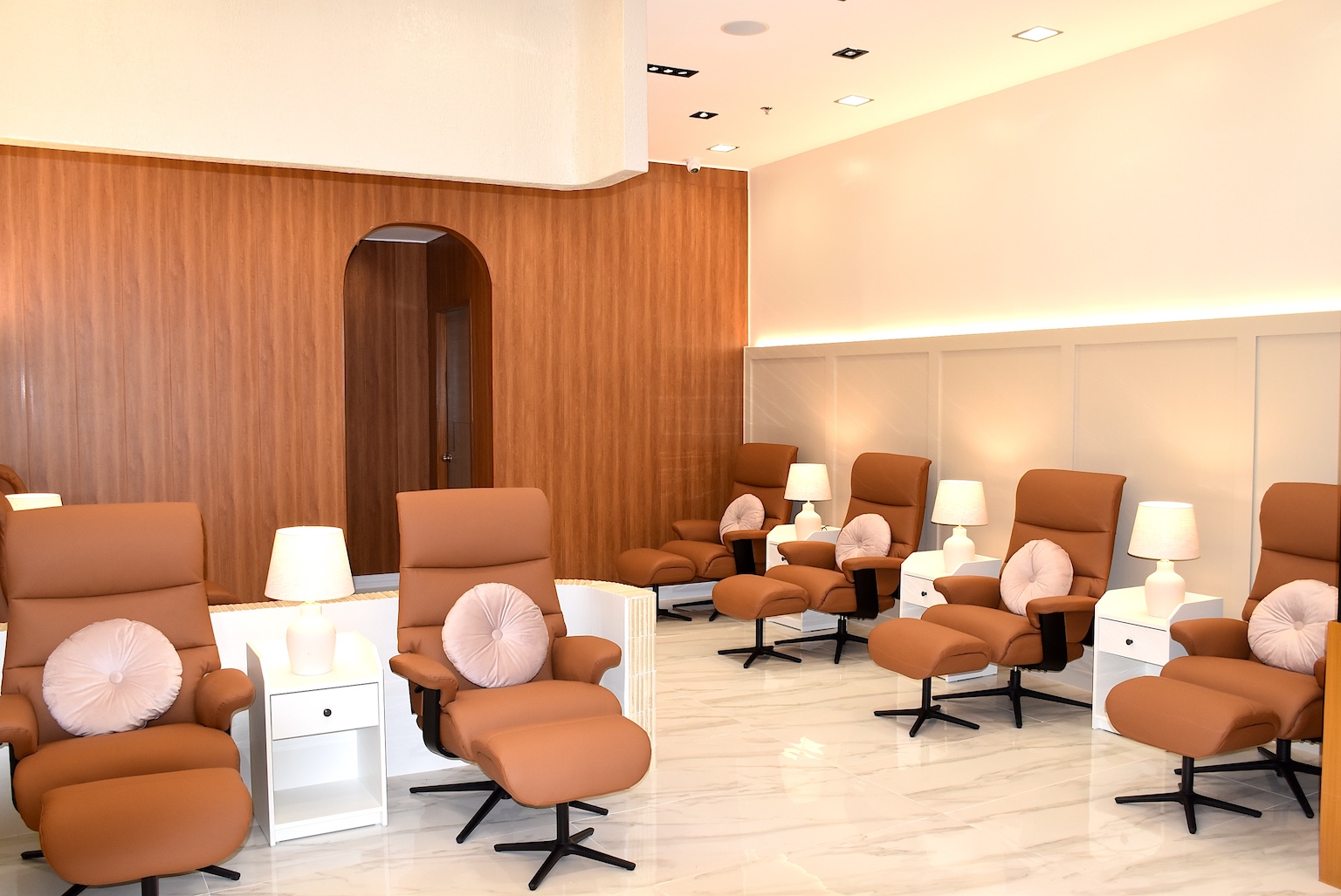 Row of brown reclining spa chairs with matching ottomans, white side tables, and lamps in a modern beige salon with wood-paneled wall behind.